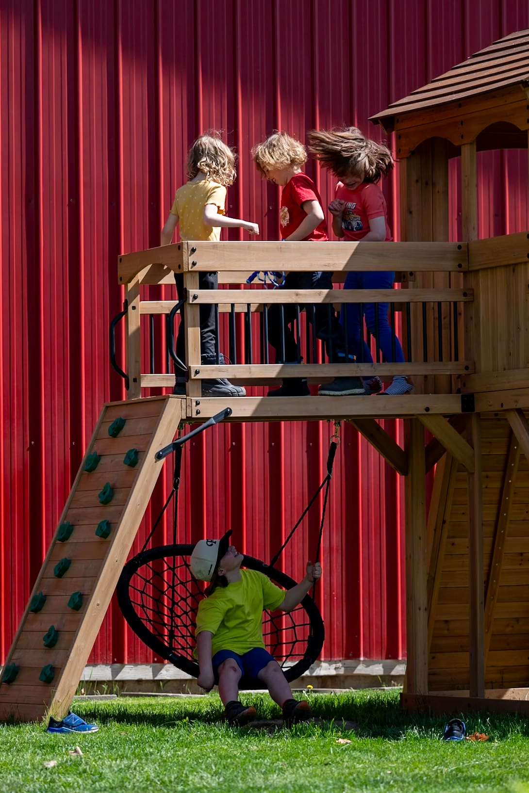 Four children are playing on a wooden playset, with two jumping on a platform and one sitting on a swing below. The background features a red wall.