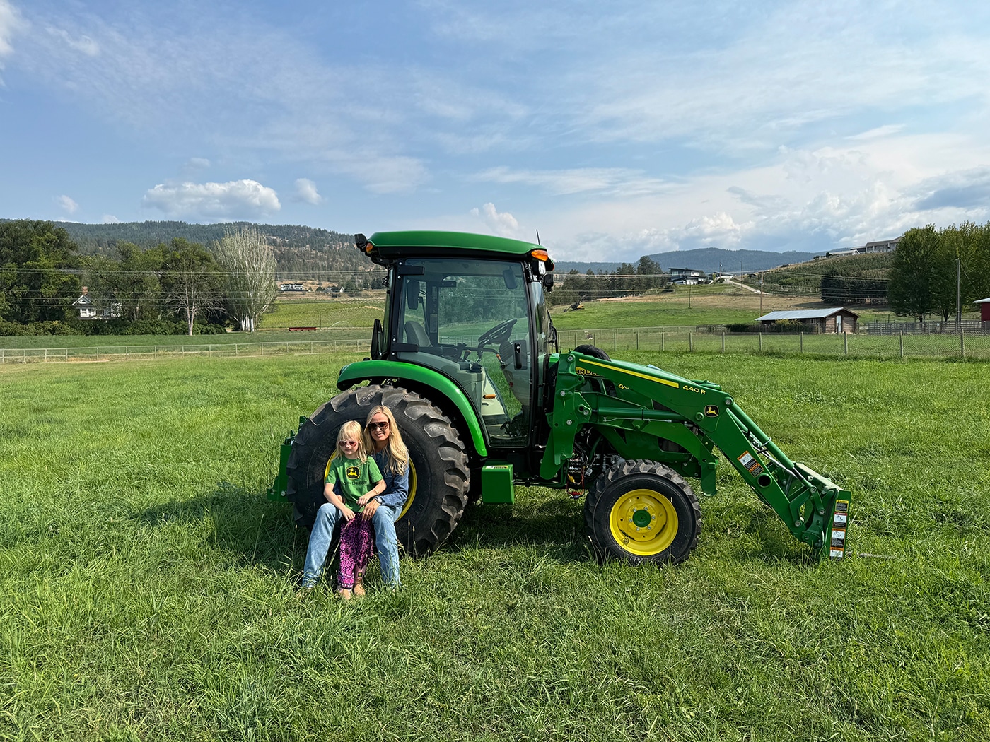 A woman and a child sit on the grass next to a green tractor in a field, with hills and buildings in the background.