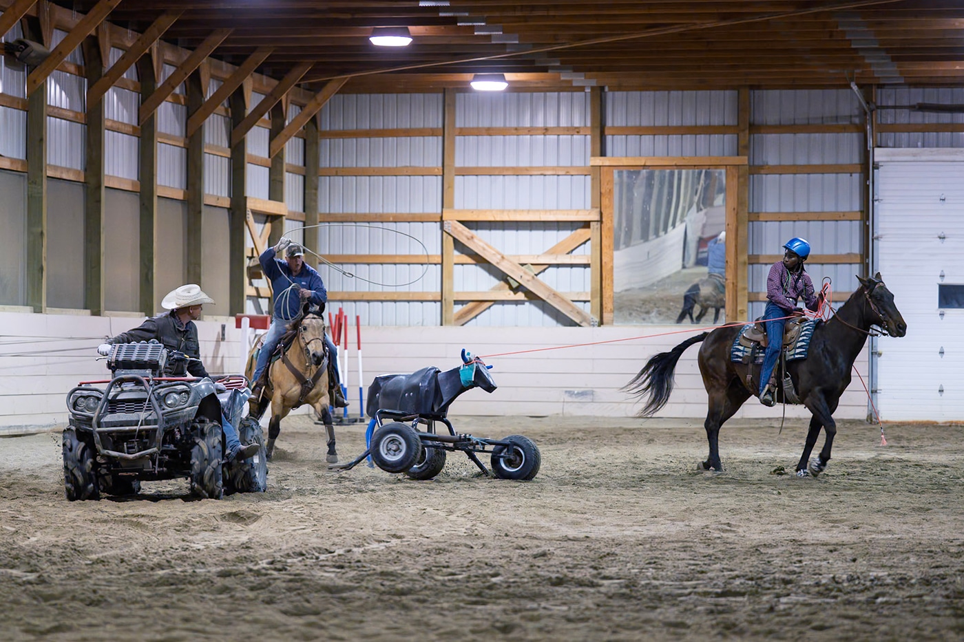 A riding arena scene with two people on horses, one using a lasso, and a third person on an all-terrain vehicle (ATV) near a training apparatus resembling a small horse.