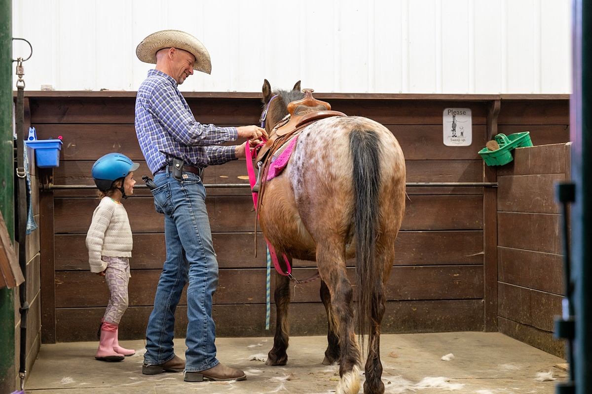 A man in a cowboy hat is adjusting a saddle on a horse while a young girl with a blue helmet watches nearby. They are in a barn setting.