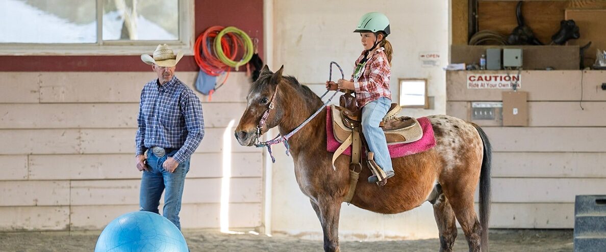 A girl wearing a helmet sits on a saddle of a horse while a man watches nearby in an indoor riding arena.
