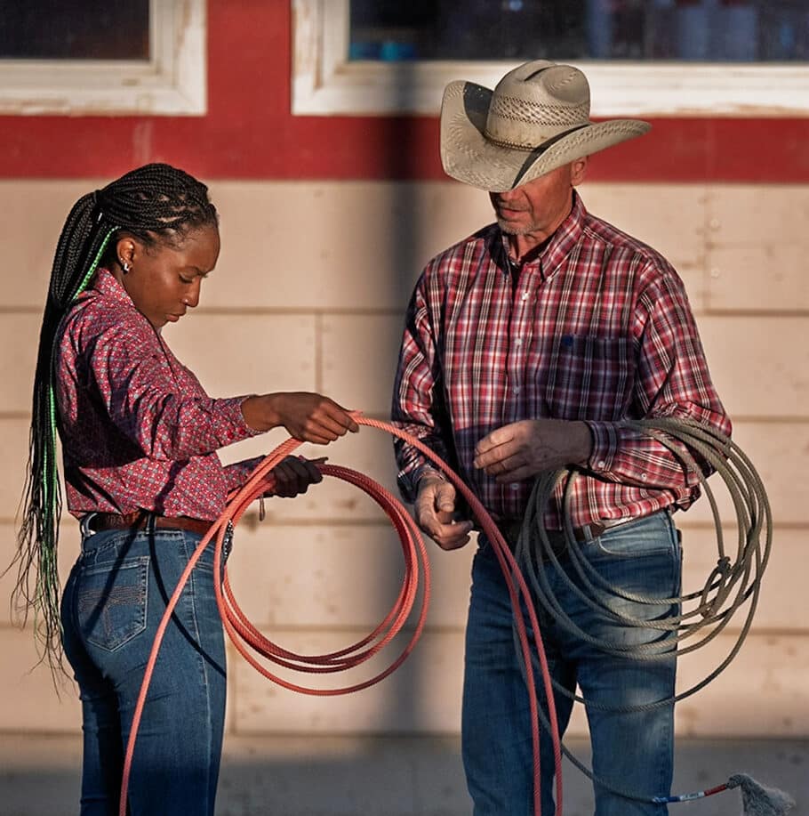 A young woman and an older man are handling ropes outside a building, with the man wearing a cowboy hat and a plaid shirt.