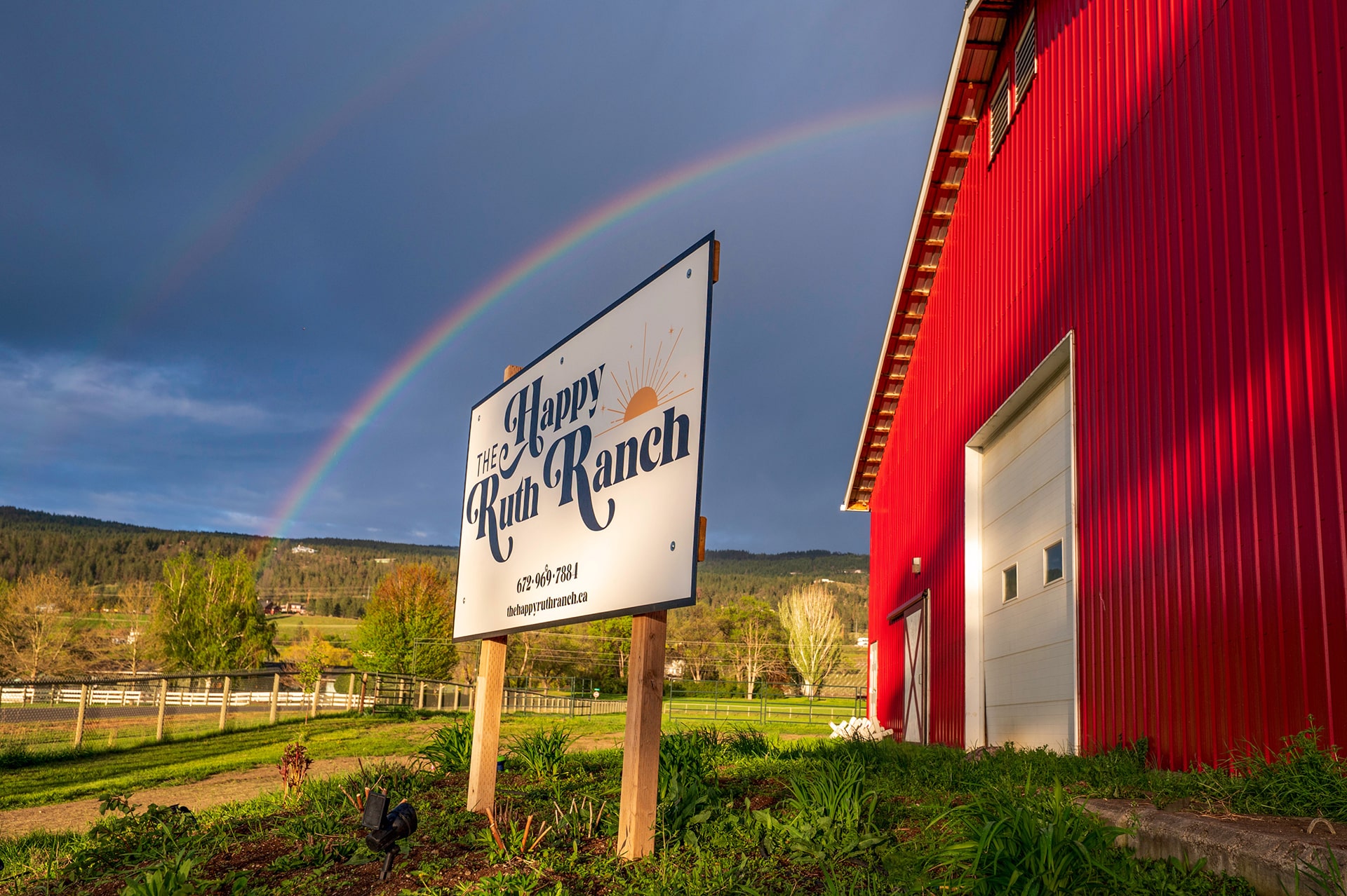 A sign for "The Happy Ruth Ranch" in front of a red barn, with a rainbow in the background and greenery surrounding the area.