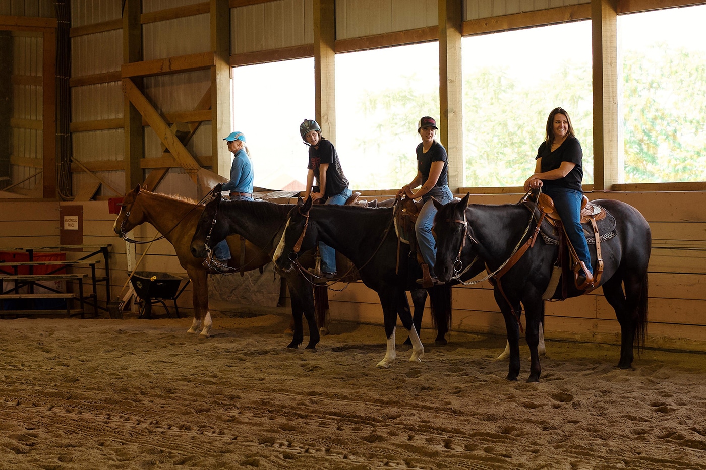 Four riders are on horseback inside an indoor arena, groomed sand floor, with large windows showing greenery outside.