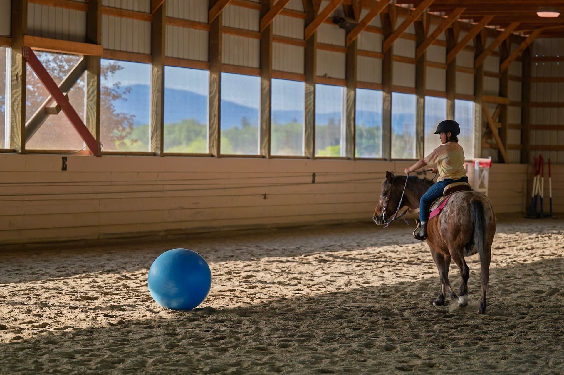 A child riding a horse in an indoor arena, near a large blue ball on the ground.