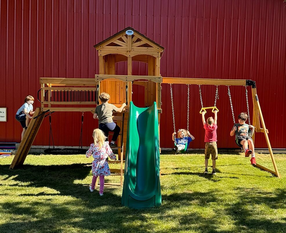 A wooden playground structure with a slide, climbing area, and swings, featuring several children playing.